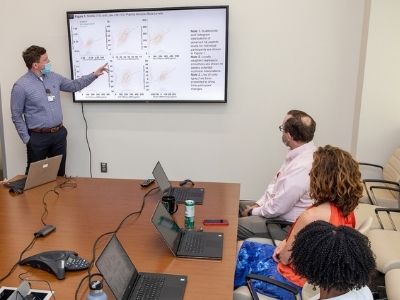 A group of researchers seated at a table listen and watch as another researcher presents findings next to a white board.